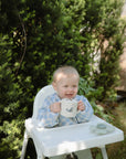 A toddler in a blue and white checkered shirt smiles outdoors, holding a mushie Snack Cup. The spill-proof silicone cup is gripped in both hands, with green bushes and sunlight brightening the background.