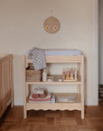 A light wooden changing table with a blue mat holds baby supplies like the mushie Organic Cotton Muslin Swaddle Blanket and toys, next to a crib. Above is a round wall decor with a smiling face. The room features parquet flooring and light walls.