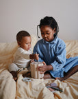 A young girl and a baby sit on a yellow bed, playing with the mushie Coin & Tube Sorting Set, as the girl helps the baby develop fine motor skills by guiding a piece into the educational toy.