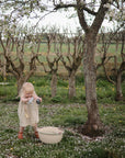 A young child in a checkered dress stands under a blossoming tree with a watering can, evoking the nurturing care behind the FRIGG Butterfly Anatomical Silicone Pacifier 2-Pack (6-18 Months)—promoting healthy baby oral development.