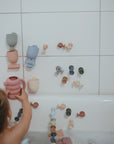 A young child delights in sensory play at bath time, stacking pastel silicone cups and sticking suction toys from the mushie Pour and Play Bath Set onto the tiled wall above the tub.