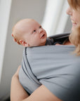 A smiling woman holds her baby close in a mushie Baby Wrap, while soft natural light fills the scene and the baby gazes up at her with curiosity.