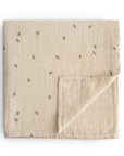 The mushie Organic Cotton Muslin Swaddle Blanket in Bees is shown folded on a white background, featuring a subtle grid texture with yellow floral embroidery and one corner turned up to display the underside.