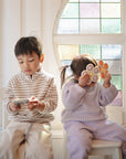 Two young children sit on white chairs by a colorful stained-glass window. The boy on the left looks at a smartphone, while the girl on the right holds up a mushie Flower Press Toy for sensory exploration.