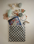 A checkered pouch holds kids’ toys, a book, bottle, stackers, silicone teethers, and a spoon—ideal for diaper bag storage with mushie Water Resistant Wet Bag in soft pastel shades.
