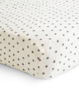 A close-up of a neatly made bed with the mushie Extra Soft Muslin Crib Sheet, featuring breathable baby bedding in white with evenly spaced small black floral designs.