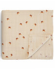 The mushie Organic Cotton Muslin Swaddle Blanket in the Butterflies design is shown folded on a white background, featuring a beige fabric with a brown and orange floral pattern and one corner slightly turned up.