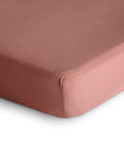 Close-up of the mushie Mini Muslin Crib Sheet in mauve, made from 100% cotton muslin, neatly fitted over a mattress corner against a white background—ideal as a soft, breathable crib sheet.