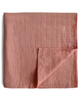A folded mushie Organic Cotton Muslin Swaddle Blanket in Cedar, featuring a subtle grid pattern. One corner is turned up to show the underside. The soft, lightweight blanket is displayed on a white background.