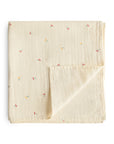The mushie Organic Cotton Muslin Swaddle Blanket in Cherries features a folded, cream base with a striped texture and scattered red, yellow, and blue floral patterns. One corner is flipped to show its soft organic cotton underside.