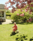 A child rides a mushie Mini Toddler Bike on a sunny lawn under pink cherry blossoms.