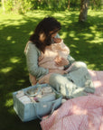 Woman on grass breastfeeding near a picnic blanket and a mushie Diaper Caddy.