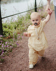 A toddler holds an adult’s hand while walking on a gravel path, using a FRIGG Daisy Natural Rubber Pacifier. She's dressed in yellow checks, with purple flowers and water visible in the background.