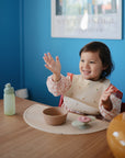 A young child in a bib sits at a wooden table, smiling and raising their hands. On the table is a mushie Silicone Suction Bowl—ideal for baby feeding time—along with a plate of food and a water bottle, against a blue wall with framed art.