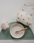 The mushie Silicone Baby Bib, cream-colored with blue whale designs, comes in a baby feeding set with a soft beige training cup and matching bowl with spoon, all on a green silicone placemat for easy cleanup.