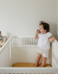 Toddler in a mushie crib with Extra Soft Muslin Crib Sheet, holding a toy and looking aside.
