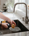 Someone washes kitchen utensils under running water in a modern sink, cleaning a grater and mushie Vehicles Mold Free Bath Play Set. Various brushes and utensils sit in a holder by the window.