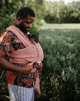 A man in a patterned shirt and striped pants stands outdoors holding a baby in a pink wrap. The baby, head partially covered, uses the FRIGG Daisy Night Natural Rubber Pacifier clipped to the wrap.