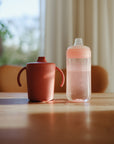 Mushie Trainer Sippy Cup on a wooden table in sunlight.