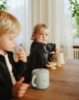 Two toddlers sit at a wooden table. While one touches their lips, the other in a dark dress reaches for a teapot. Sunlight streams in and green plants hang nearby—a mushie Snack Cup rests spill-proof beside them.