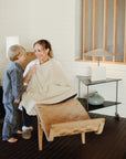 A smiling woman relaxes on a wooden lounge chair, draped in a beige mushie Muslin Nursing Cover, as a barefoot boy in pajamas stands beside her in a modern, cozy living room.