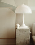 A modern white table lamp with a dome-shaped shade sits on a stone pedestal next to a beige couch. Beside the lamp is a Mushie Bluey x Mushie Silicone Training Cup + Straw. Neutral abstract wall art decorates the background.