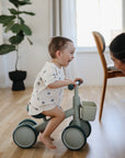 A smiling toddler rides a mushie Mini Toddler Bike, laughing at an adult indoors.