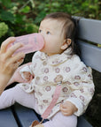 Baby on a bench, fed by an adult with a mushie Soft Spout Sippy Cup, wearing a floral jacket.