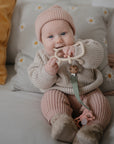 A baby in a pink knit hat and outfit sits on a couch, holding and chewing on a FRIGG Butterfly Anatomical Silicone Pacifier (6-18 Months). Pillows, including one with daisies, and a pink blanket are in the background.