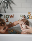Two kids play in the tub with mushie's Dino Mold Free Bath Play Set, passing a toy egg to an adult. The silicone bath toys offer safe, mold-free fun. White tiles and a faucet are in the background.