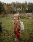 A young child with a tan backpack and rust-colored pants stands on grass near a pig, using the mushie Silicone Pacifier Clip | Hera to secure their pacifier, surrounded by trees outdoors.