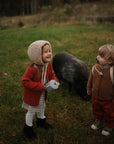 Two toddlers in hats and boots stand on grass, smiling at each other while holding mushie Snack Cups. Both cups are spill-proof and made of silicone. A black pig grazes nearby, adding to the cozy autumn outdoor scene.