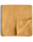 The mushie Organic Cotton Muslin Swaddle Blanket in Fall Yellow is neatly folded with one corner turned up, showing subtle vertical stitching. The blanket is displayed on a white background.