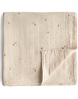 The mushie Organic Cotton Muslin Swaddle Blanket in Falling Stars features light beige organic cotton with a subtle grid and small brown stars and shooting star designs, neatly folded to display the soft underside.