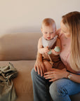 A mother sits on a beige sofa holding her baby, who plays with a sage green silicone teether on a wooden bead chain. Beside them, the Mushie Ribbed Baby Blanket is neatly folded.