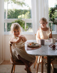 Two young children in light dresses sit at a sunlit dining table with potted plants, enjoying their toddler feeding essentials from the mushie Deluxe Pink Flowers/Blush Feeding Kit.