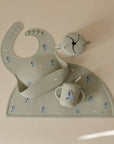A mushie Bluey x Mushie Snack Cup, pale green bib, straw cup, and placemat with white stars and cartoon dogs are arranged on a beige surface. The feeding essentials showcase Bluey and white dot accents.