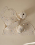 The mushie Bluey x Mushie Silicone Baby Bib, in light colors with playful dog and heart patterns inspired by Bluey, is shown neatly arranged on a cream background.