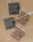 Four neatly folded mushie silicone bibs in gray, sage green, blush pink, and blush with prints are shown in packaging on a beige surface. Each set comes with a matching mushie Water Resistant Wet Bag for easy diaper bag storage.