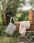 A vintage brown stroller sits on grass near bushes. A checkered bag and a mushie Water Resistant Wet Bag hang from the handle, while a striped blanket with a pacifier rests on the side.