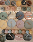 A flat lay of mushie Round Dinnerware Plates in pastel pink, yellow, blue, green, and beige—non-toxic and dishwasher safe—arranged neatly on a light surface.