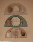 Three mushie BPA-free children's square dinnerware sets: beige set (cup, bowl, spoon, toothbrush), green set (plate, cup, spoon), and pink set (square plates, cup, fork, spoon) on semicircular placemats.