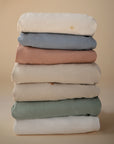 A stack of six mushie Stretchy MINI Crib Sheets in white, green, beige, pink, blue, and cream rests against a neutral background. The top sheet is breathable organic cotton and features an embroidered sun symbol.