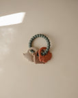 The mushie Dino Teething Ring features three textured animal-shaped charms in neutral tones on a beaded gray silicone ring, displayed on a light beige surface with gentle sunlight and shadows.