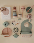 A selection of pastel mushie baby items, including non-toxic silicone bibs, plates, spoons, stacking rings toy, teether, bowls, measuring cups, rattle, and boxed muslin cloth—neatly arranged on a light beige surface.