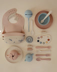 A flat lay of pastel-colored baby feeding items for toddlers, featuring mushie's Snack Cup along with silicone bibs, bowls, plates, utensils, a rattle, a cup with animal ears, and a paint palette-shaped dish on a cream surface.