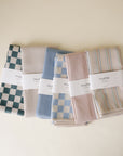 Four folded Mushie muslin crib sheets—green and white checks, solid blue, blue checks, and pastel stripes—are lined up on a neutral surface next to the Mushie Splat Mat. Each sheet features a Mushie label.