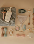 Flat lay of Mushie baby care items on beige, featuring a folded towel, lotion bottle, pacifiers, Organic Cotton Bath Mitt for sensitive skin, teethers, soft dumbbells, headband, sippy cup, and pastel clip-on teether holders.
