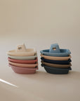 Two stacks of mushie Bath Boats in muted shades—pink, beige, blue, and navy—are arranged side by side. Each stackable boat nests neatly and is made from non-toxic plastic for safe water play.