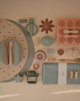 A neatly arranged set of pastel mushie baby items—including spoons, plates, bibs, pacifier clip, toys, cloth napkins, and a Finger Toothbrush for baby teeth cleaning—displayed on a light background.